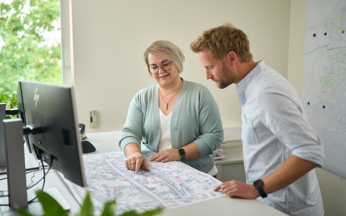 Zwei Personen stehen in einem Büro an einem Tisch und betrachten gemeinsam einen großen Stadtplan. Die Frau zeigt auf eine Stelle auf dem Plan, während der Mann aufmerksam zuhört. Im Hintergrund sind ein Computer, ein Fenster mit Blick ins Grüne und eine Karte an der Wand zu sehen.