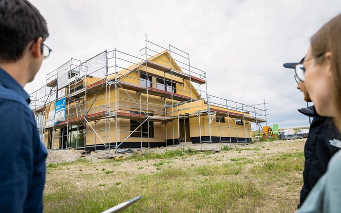 Mehrere Personen betrachten ein Einfamilienhaus im Rohbau, das von einem Baugerüst umgeben ist.
