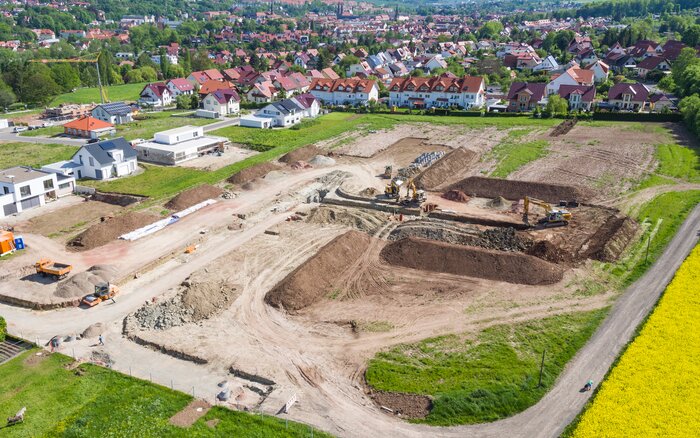 Luftaufnahme einer Baustelle am Rande eines Wohngebiets mit mehreren Baggern und Erdarbeiten. Im Vordergrund sind Erdhügel, Gräben und Baumaschinen zu sehen, im Hintergrund stehen bereits fertige Wohnhäuser mit roten Dächern und gepflegten Gärten.