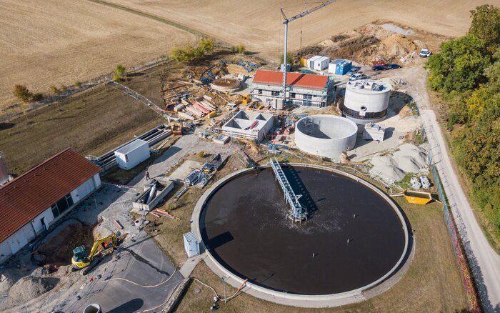 Luftaufnahme einer Kläranlage im ländlichen Raum, die sich im Ausbau befindet. Zu sehen sind mehrere runde Becken, ein zentrales Betriebsgebäude mit rotem Dach, Baugeräte, Baumaterialien und ein Kran. Die Anlage ist von Feldern und einem kleinen Waldstück umgeben.