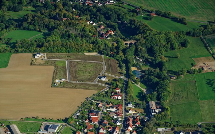 Luftaufnahme des neuen Wohngebiets. Im unteren Bereich des Bildes befinden sich bereits errichtete Häuser, während angrenzende Flächen noch unbebaut sind. Das Gebiet ist von Feldern und Grünflächen umgeben, eine Straße durchquert das Wohngebiet. Im oberen Bildbereich sind weitere Häuser locker zwischen bewaldeten Flächen verteilt.
