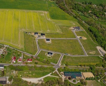 Luftaufnahme eines neu erschlossenen Wohngebiets mit Feldern, Straßen und vereinzelten Häusern. Im Vordergrund sind einige Wohnhäuser und Gärten zu sehen, während im Hintergrund große landwirtschaftliche Flächen dominieren.