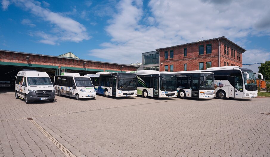 Sechs Busse und ein Van stehen vor einem Backsteingebäude bei blauem Himmel.