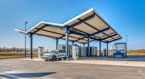 Moderner, überdachter Ladepark mit mehreren Ladesäulen und einem Elektrofahrzeug vor blauem Himmel.