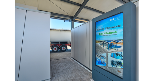 Digitale Informationstafel im überdachten Bereich des EW-Ladeparks mit Willkommensbotschaft und Bildern der Ladeinfrastruktur.