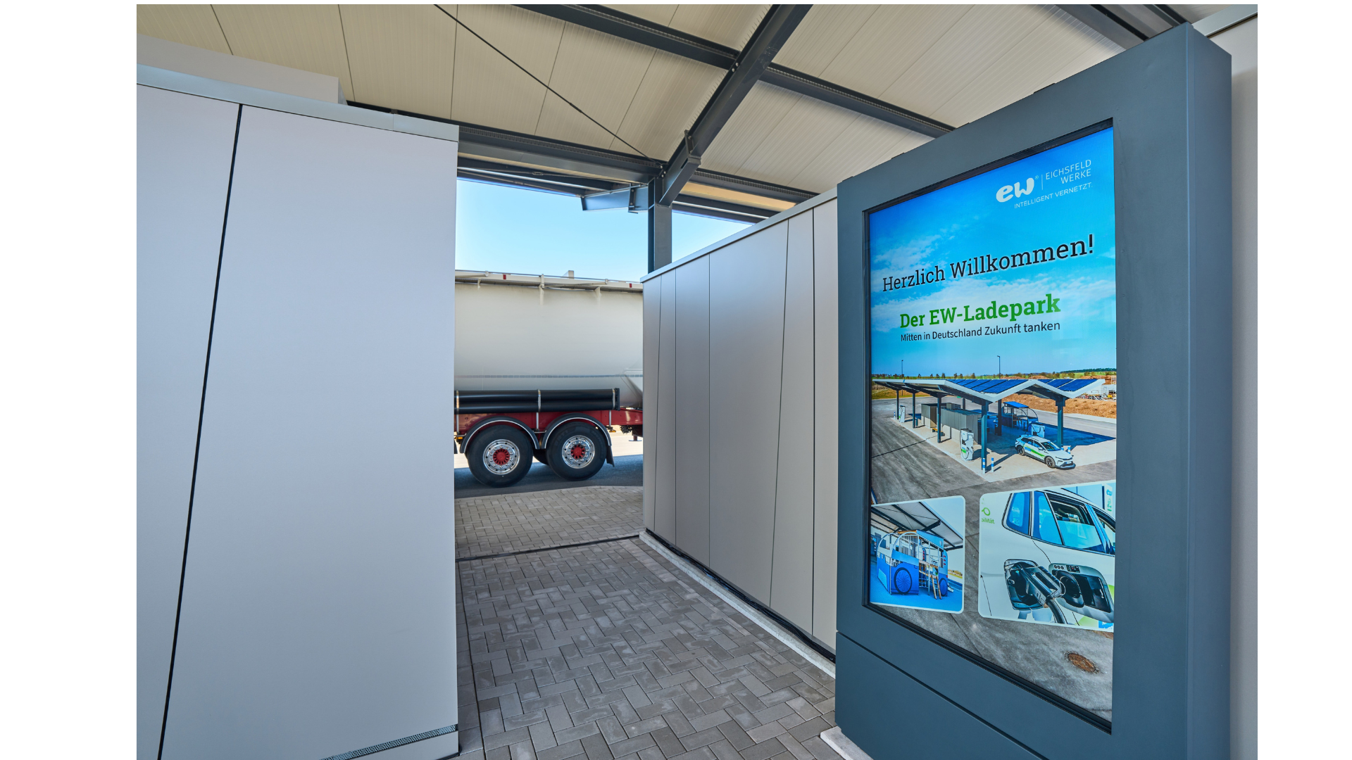 Digitale Informationstafel im überdachten Bereich des EW-Ladeparks mit Willkommensbotschaft und Bildern der Ladeinfrastruktur.