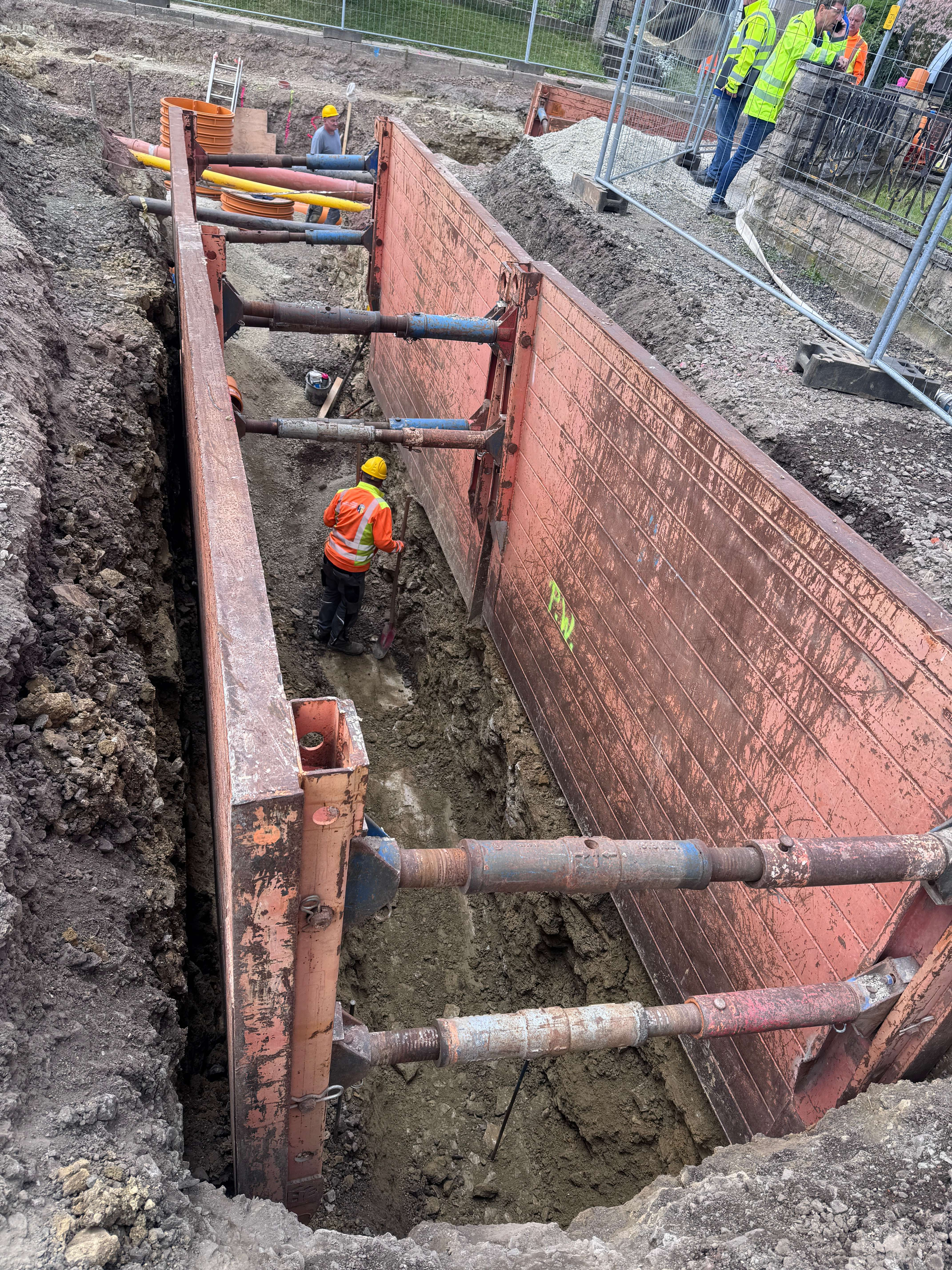 In dem mit Verbauplatten gesicherten Tiefgraben arbeiten Fachkräfte mit Schutzhelmen und Warnkleidung an der unterirdischen Infrastruktur.