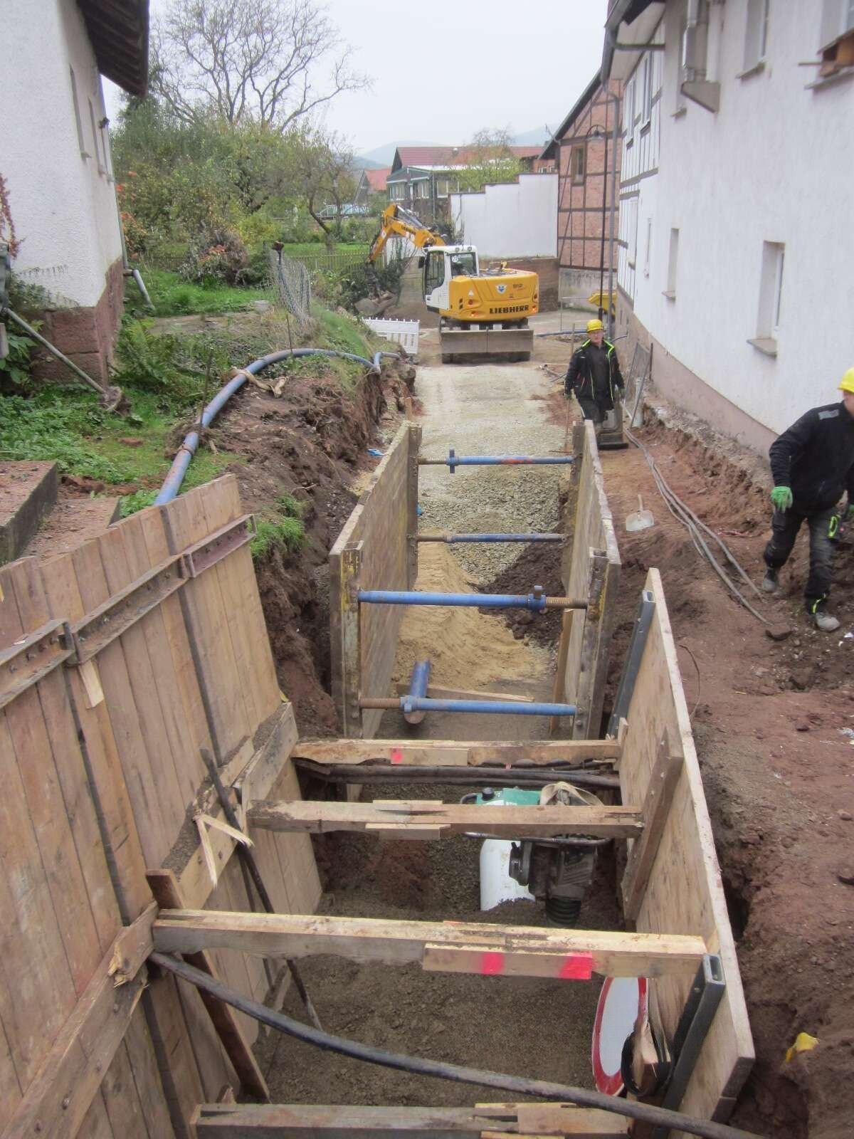 Ein mit Verbauplatten gesicherter Leitungsgraben mit Bagger und Bauarbeiter im Hintergrund.