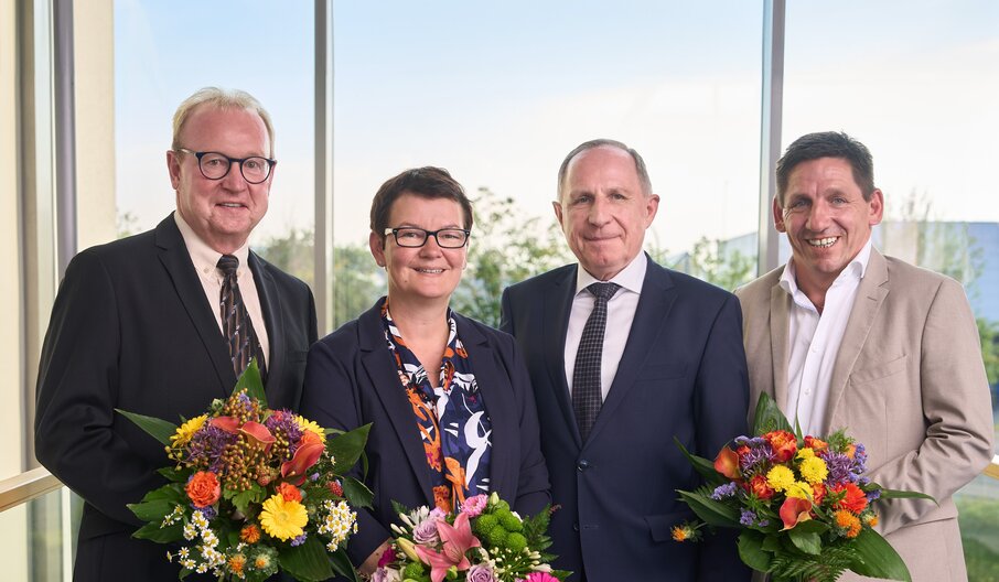 Vier Personen mit Blumensträußen vor großem Fenster mit Blick ins Grüne.
